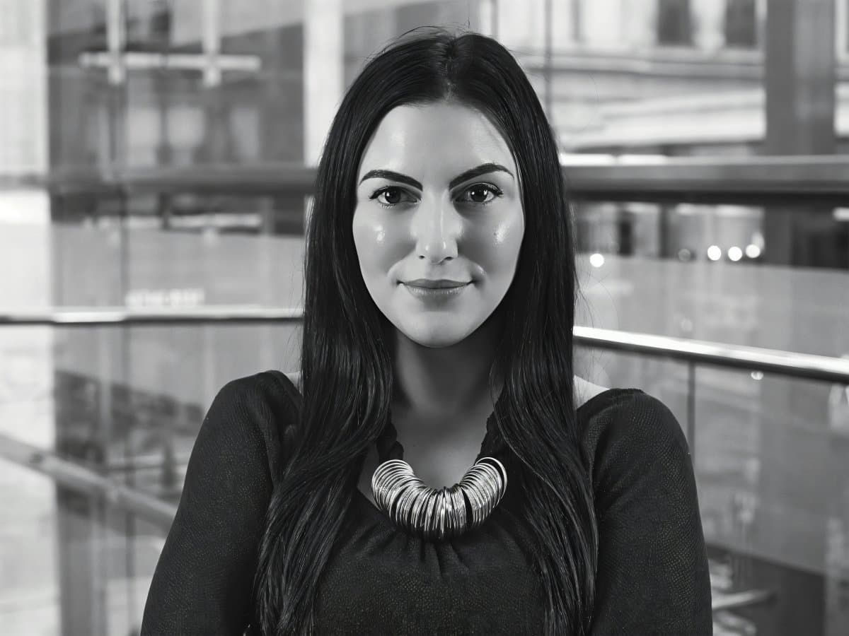 Alexandra Paras smiling in a black top and layered silver necklace against a glass office interior