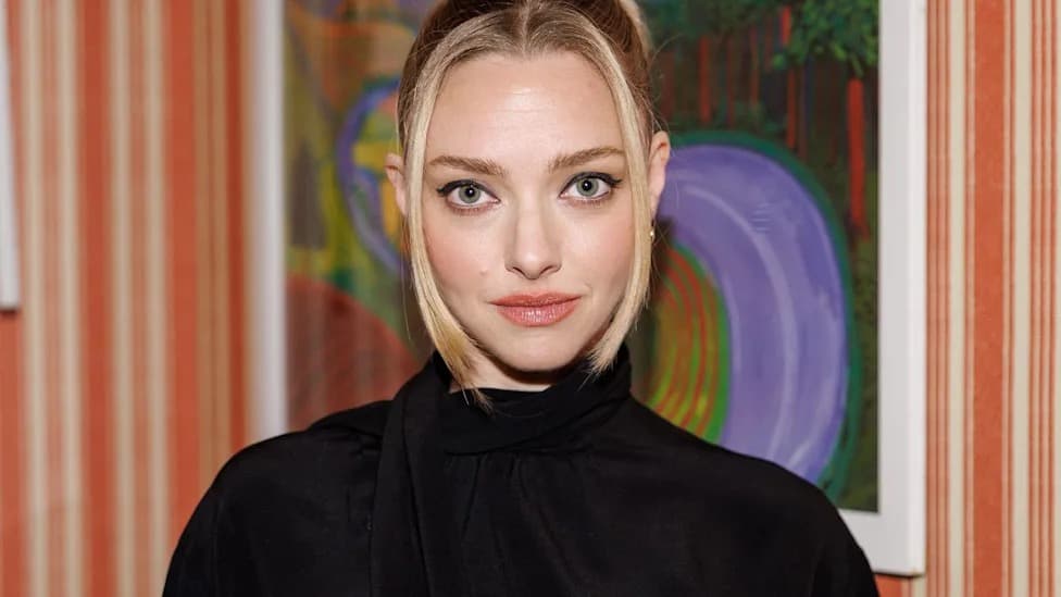 Amanda Seyfried in a black high-neck top posing in front of a colorful painting and striped wallpaper
