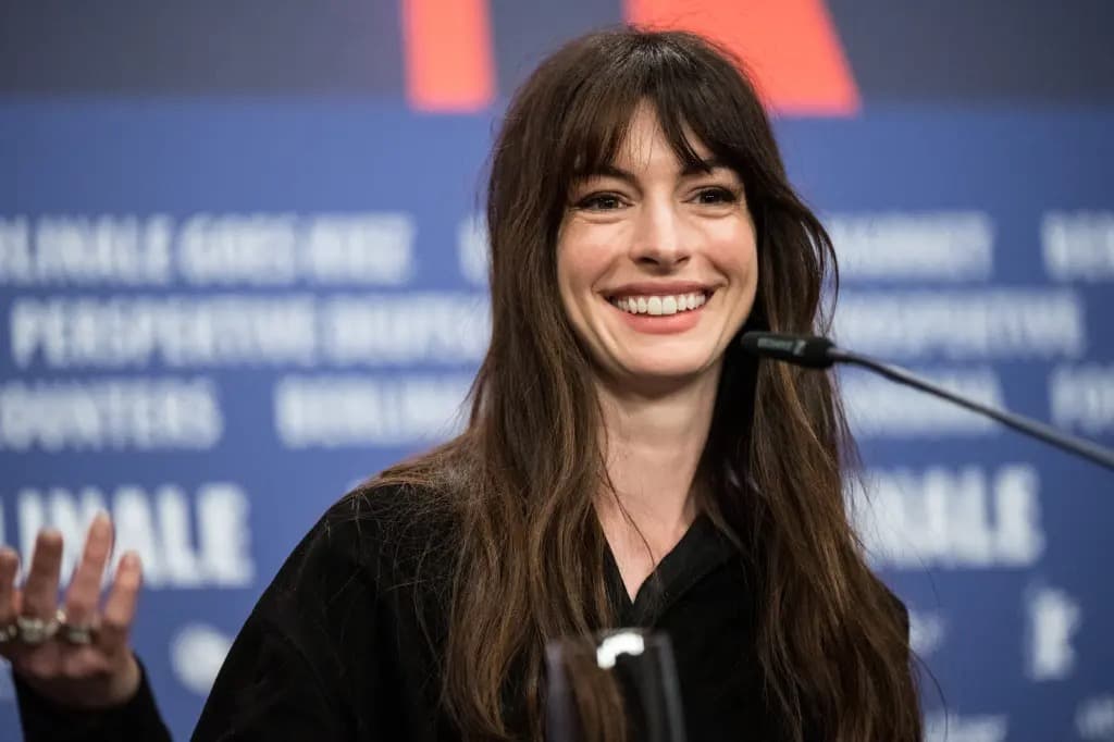 Anne Hathaway smiling and speaking at a press conference microphone in front of a blue Berlinale backdrop