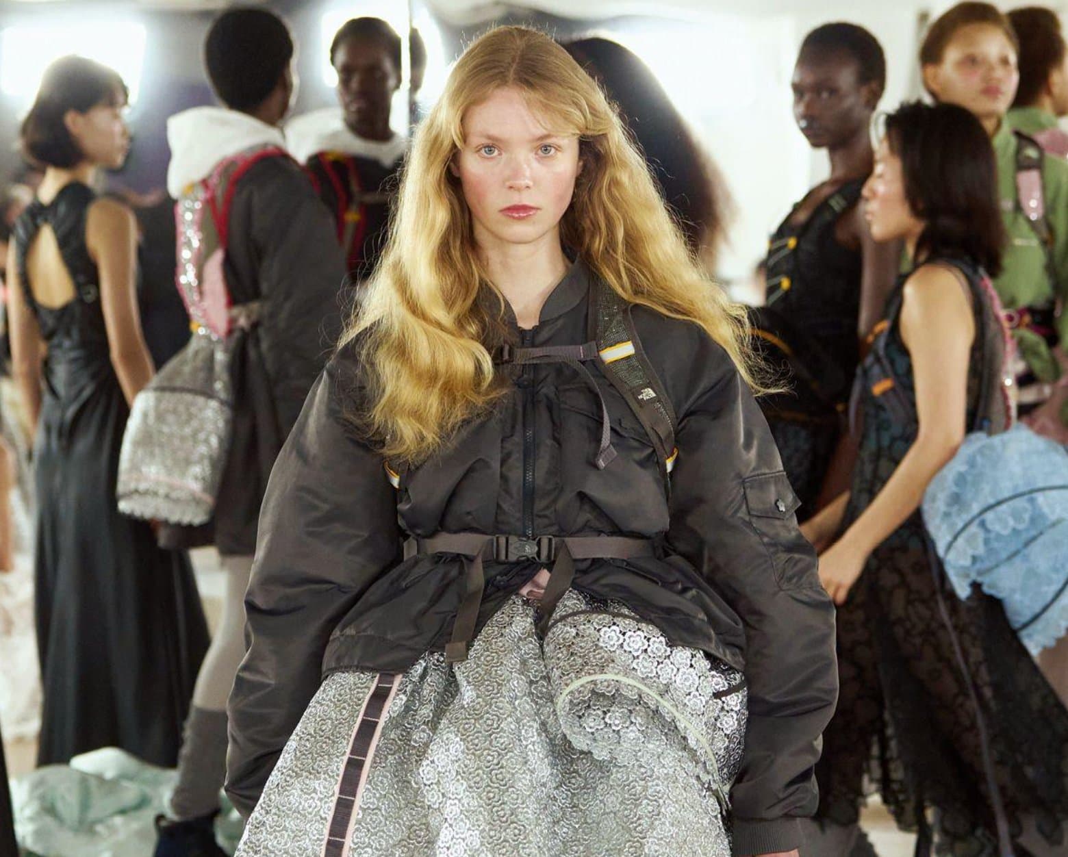 A model backstage wearing an oversized dark bomber jacket cinched with straps over a silver floral lace skirt