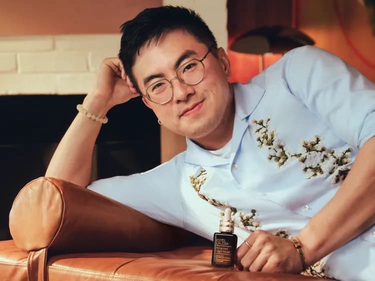 Bowen Yang relaxing on a leather couch in a floral embroidered shirt, holding a bottle of Estee Lauder Advanced Night Repair serum