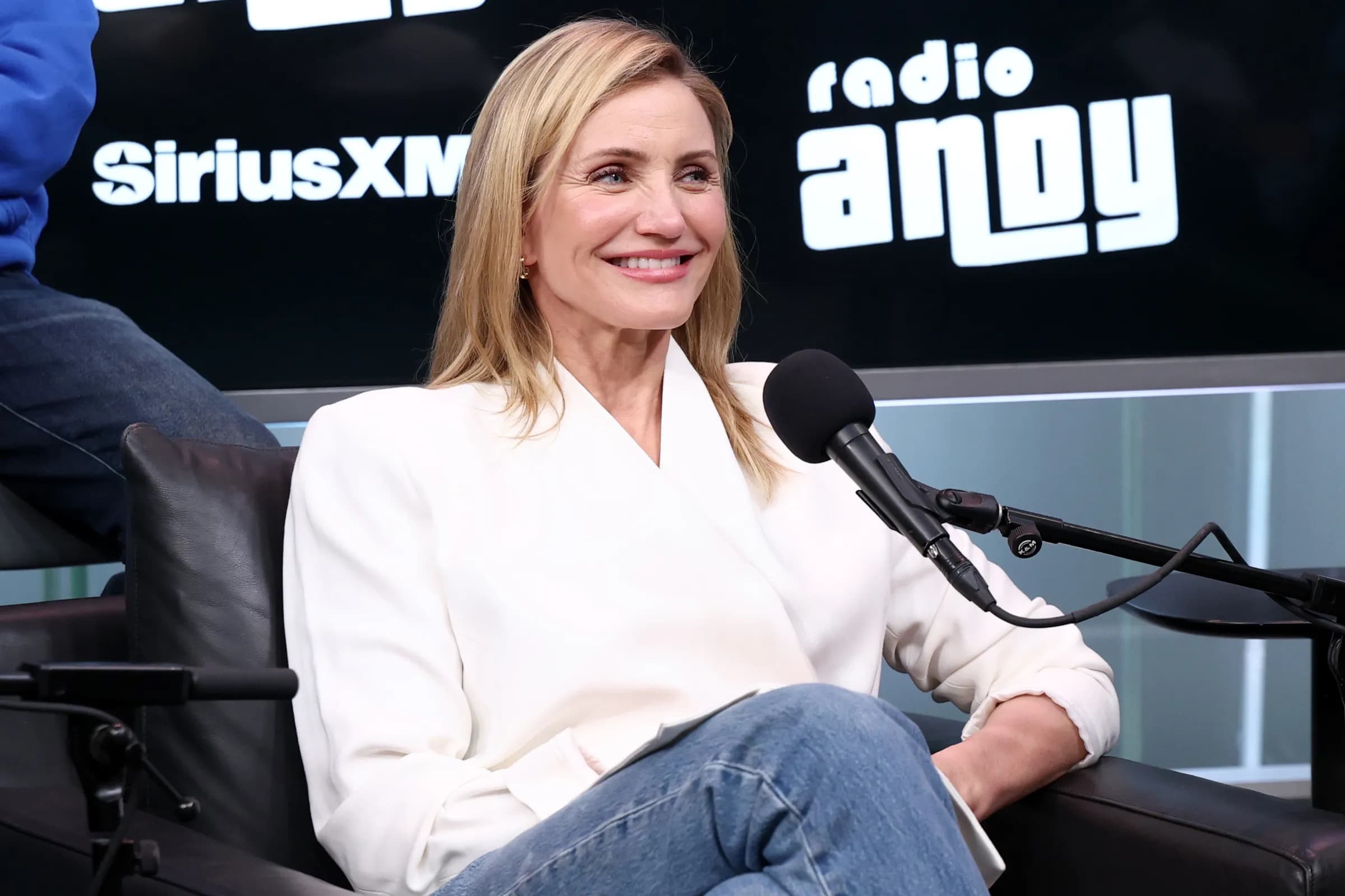 Cameron Diaz smiling while seated at a SiriusXM Radio Andy interview, wearing a white blazer and jeans