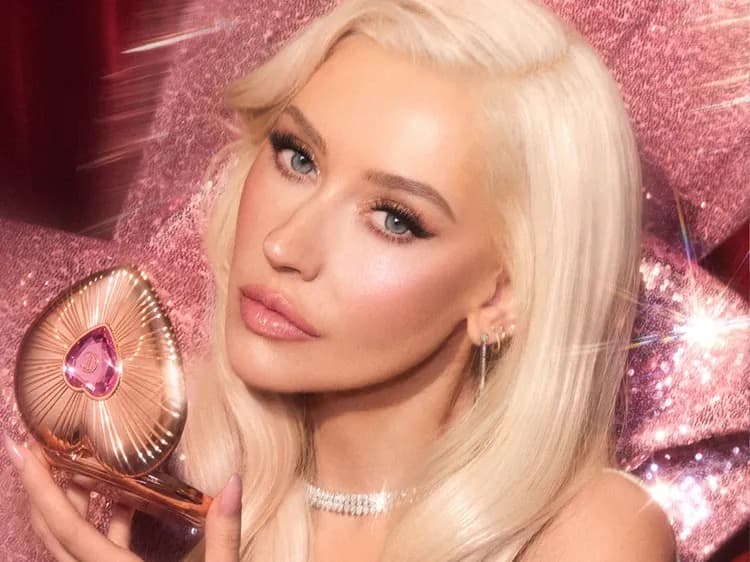 Christina Aguilera with platinum blonde hair holding a glowing heart-shaped pink compact against a sparkling pink backdrop