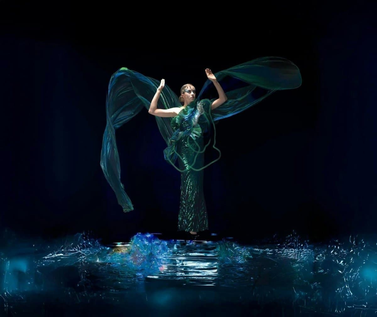 A model in a flowing green sequined gown with sheer wing-like fabric extending from her arms, standing above shattered glass-like water effects