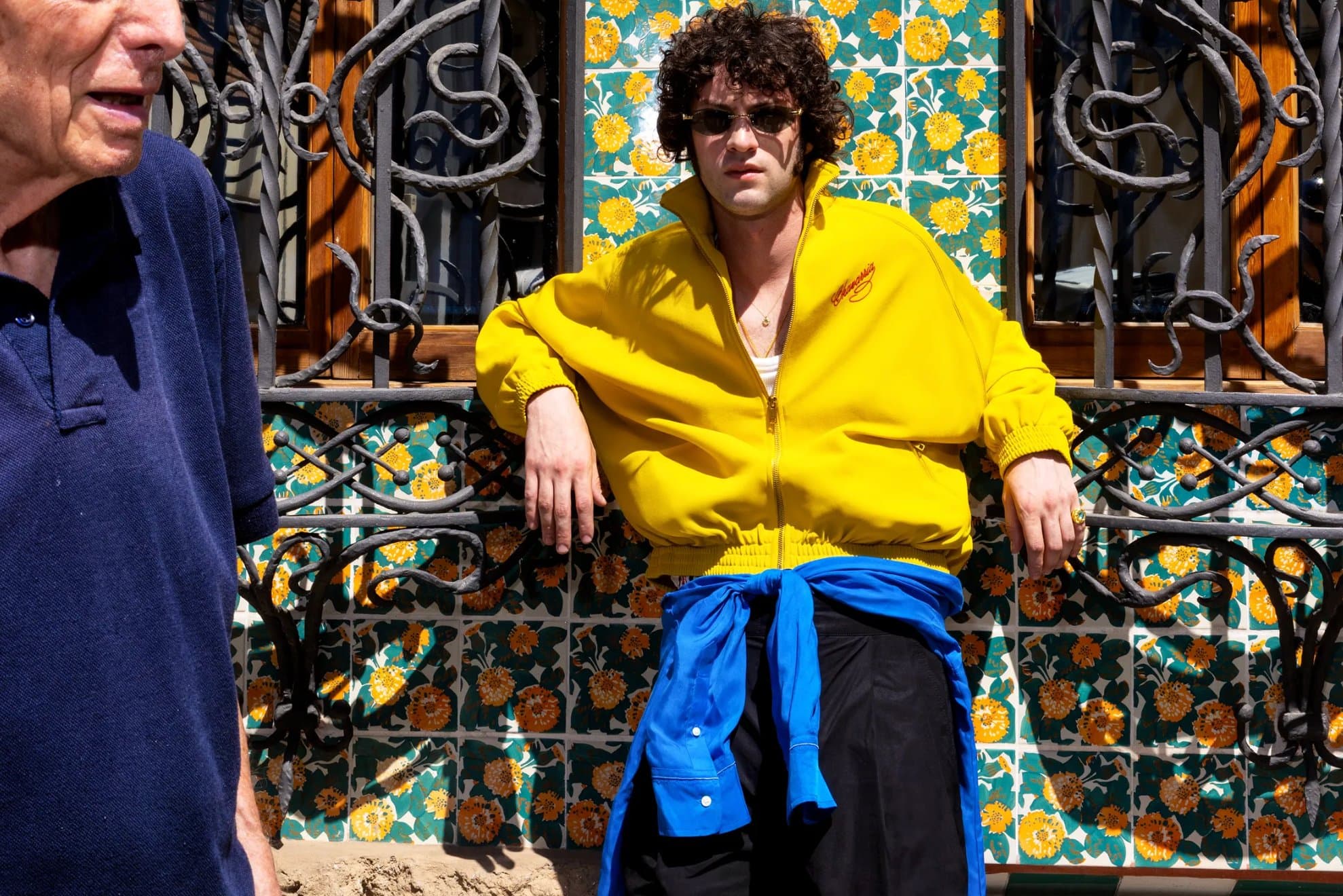 Dominic Sessa in a yellow jacket leaning against an ornate wrought-iron window grille backed by floral tiles