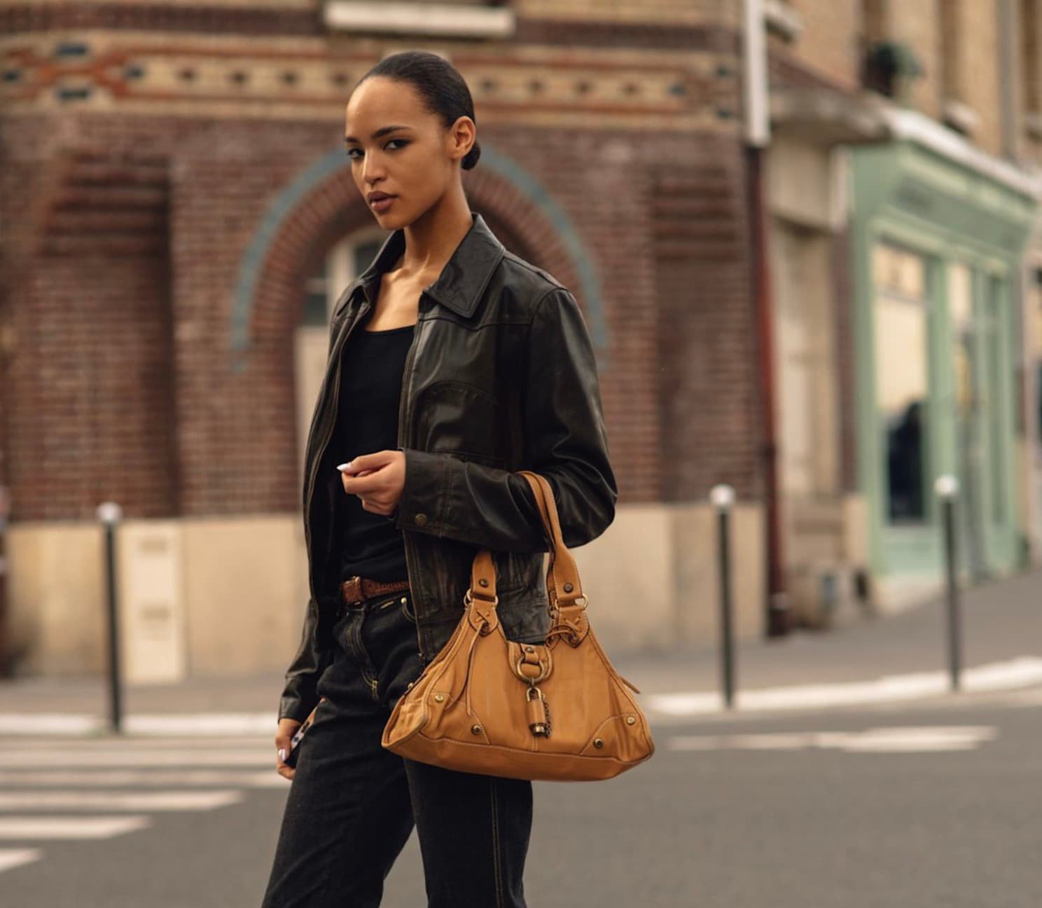 Woman on a city street wearing a black leather jacket over a black top and dark denim, carrying a tan leather handbag