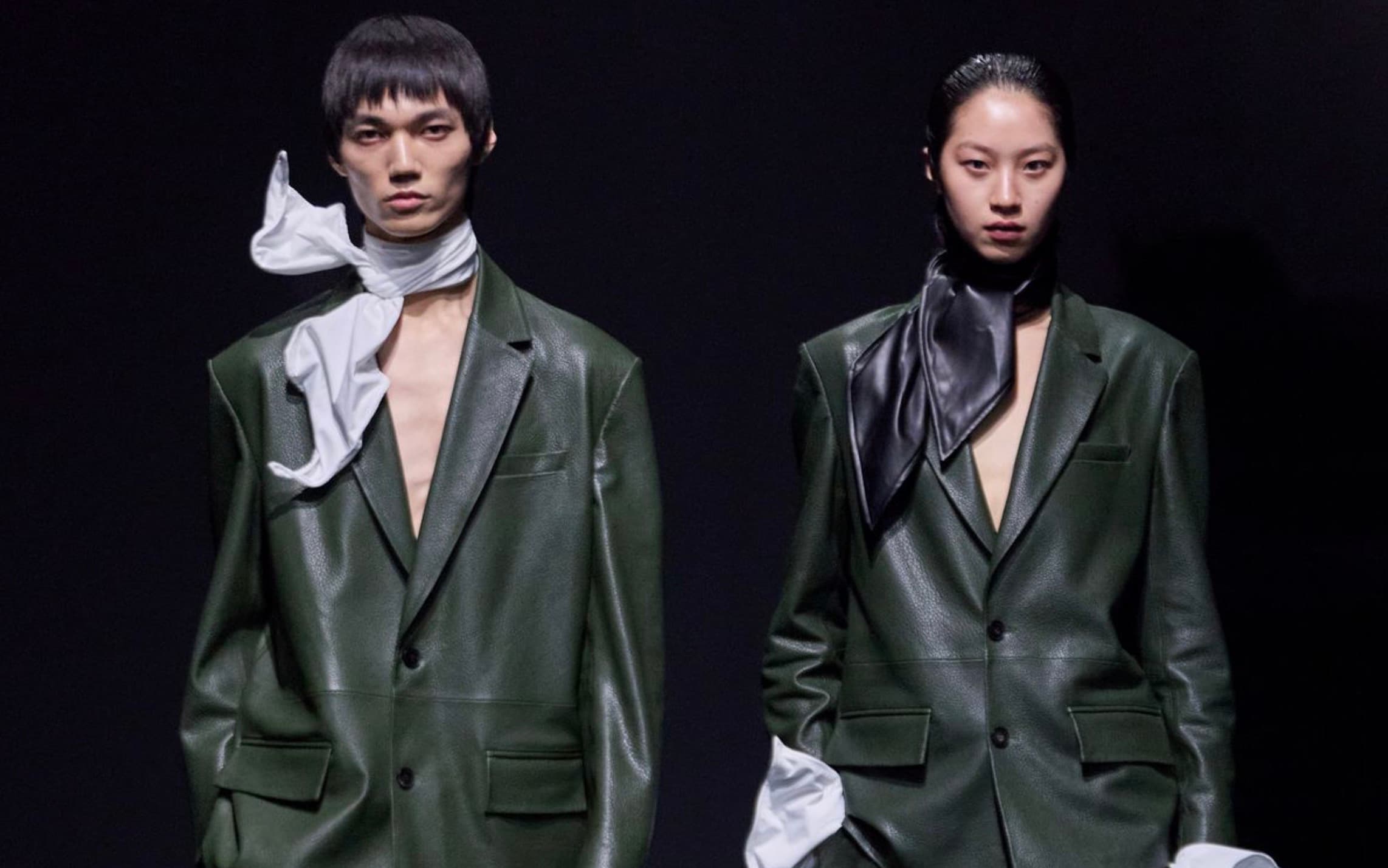 Two runway models in matching oversized olive green leather blazers, one with a white scarf and one with a black scarf