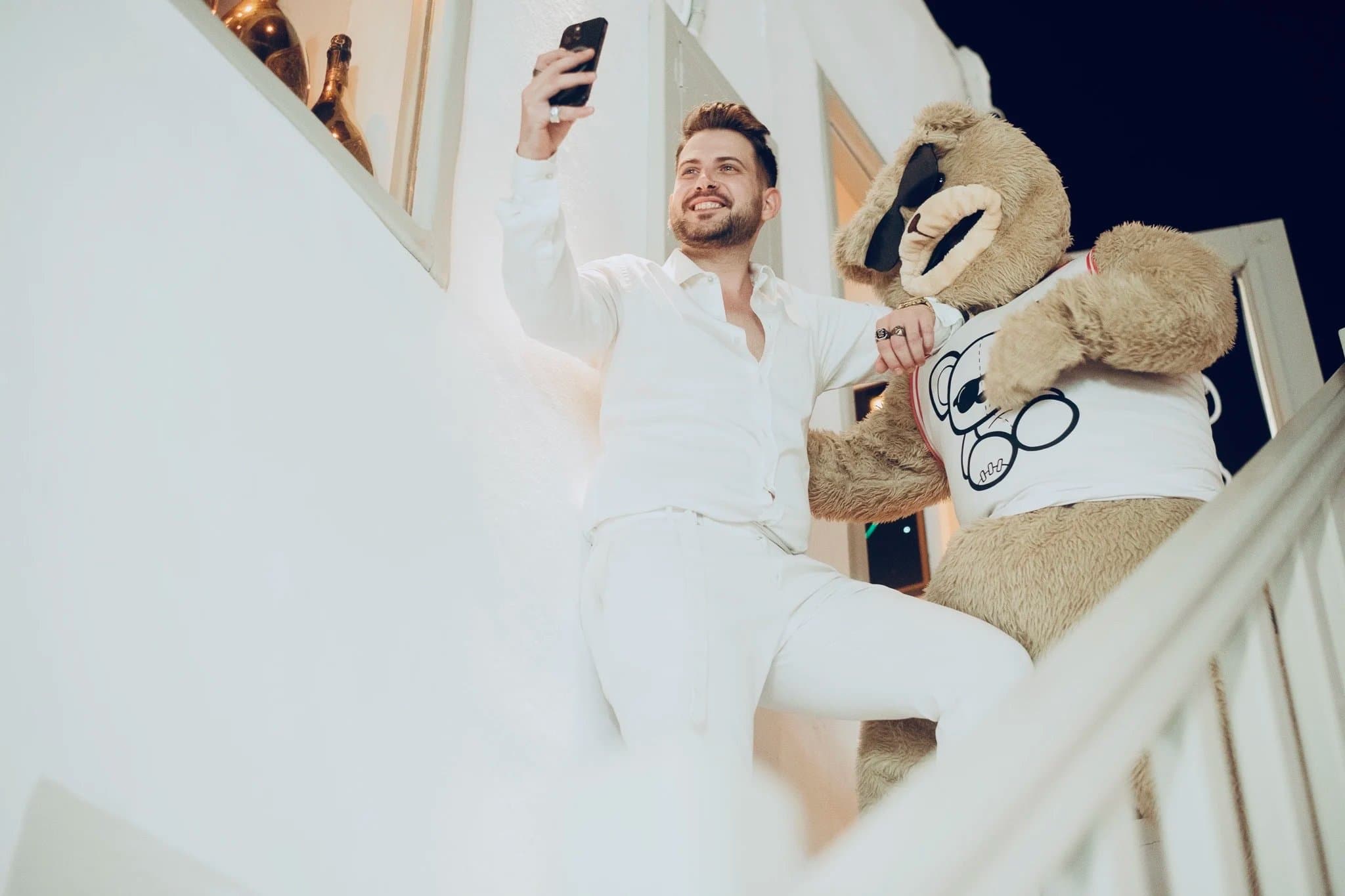 A man in white posing for a selfie with a person in a large teddy bear costume on an outdoor staircase at night