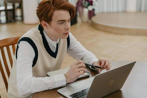 A young man with red hair taking notes at a wooden desk while working on a laptop