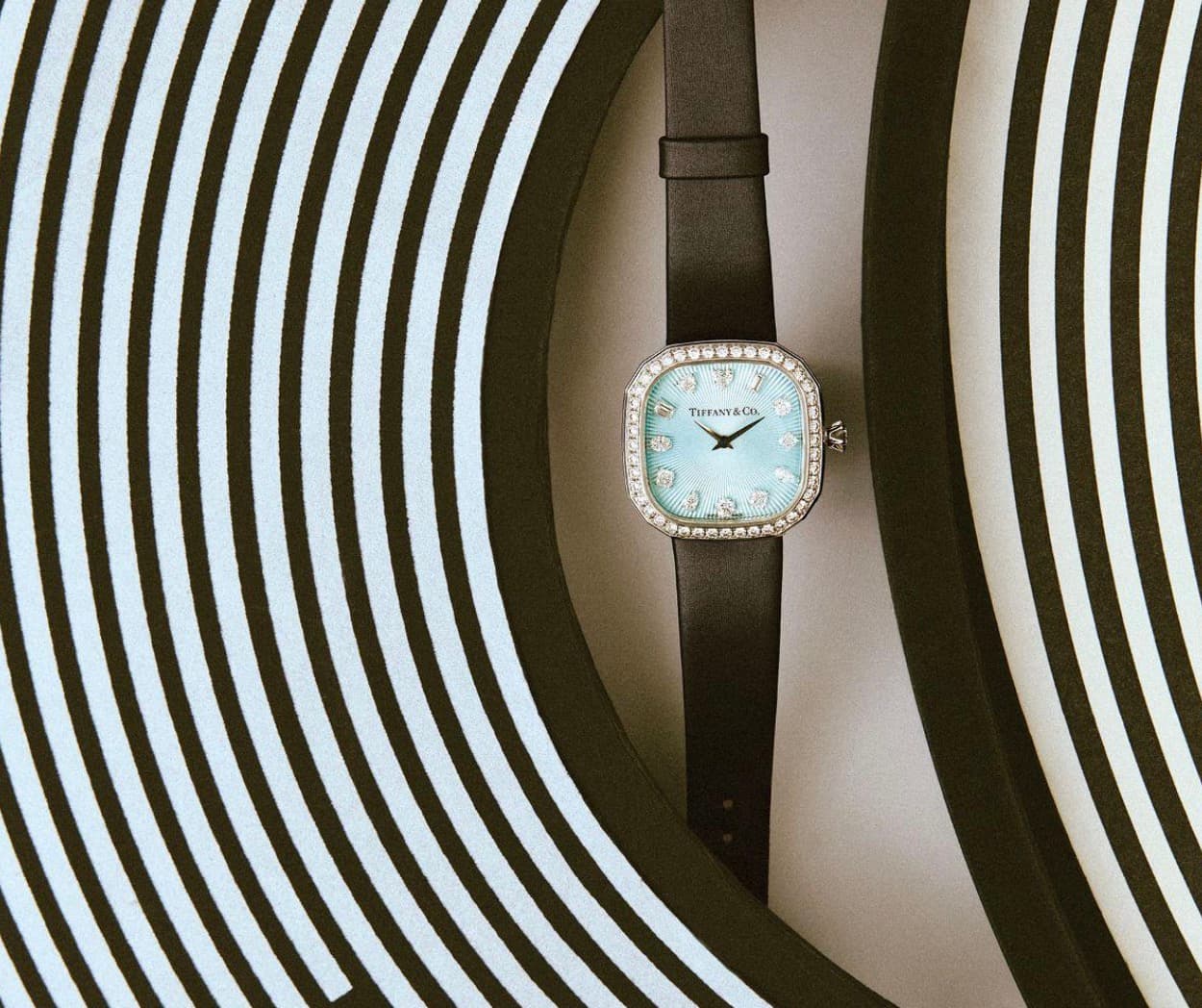 A Tiffany & Co watch with a light blue dial and diamond bezel resting against a black and white striped surface