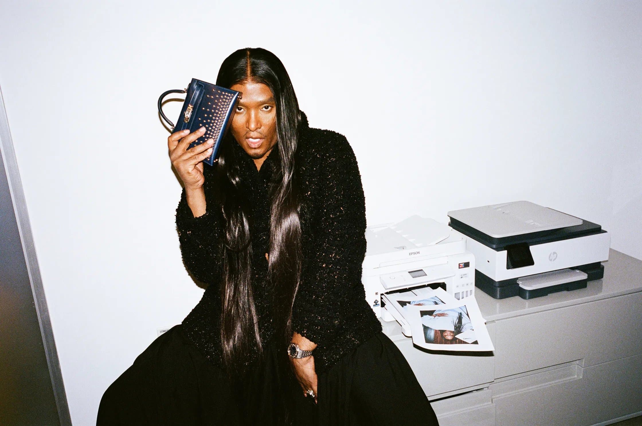 Law Roach in a black sequined jacket holding a navy studded handbag up to his face
