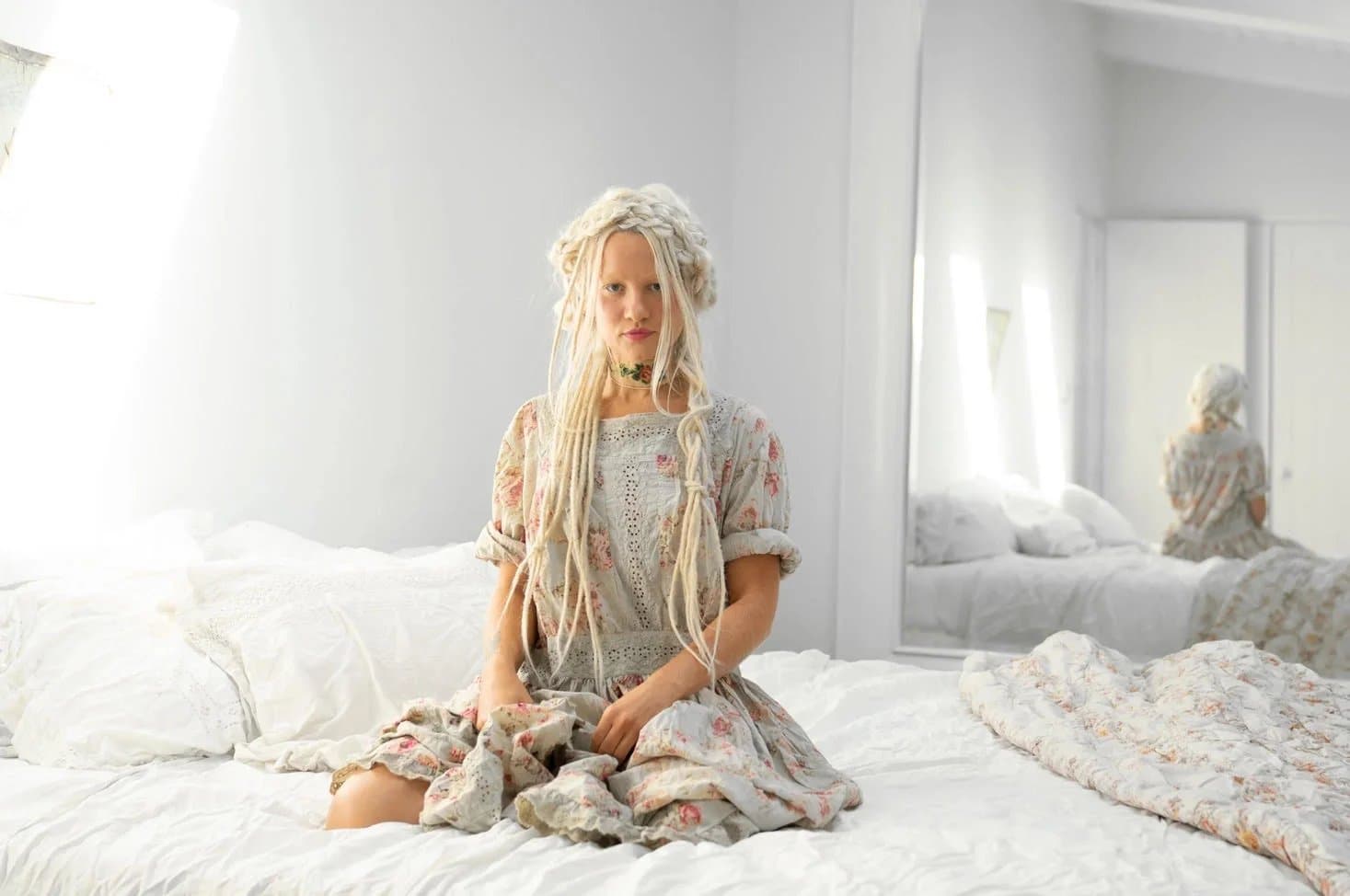 A woman with braided blonde hair sitting on a white bed wearing a floral vintage-style dress