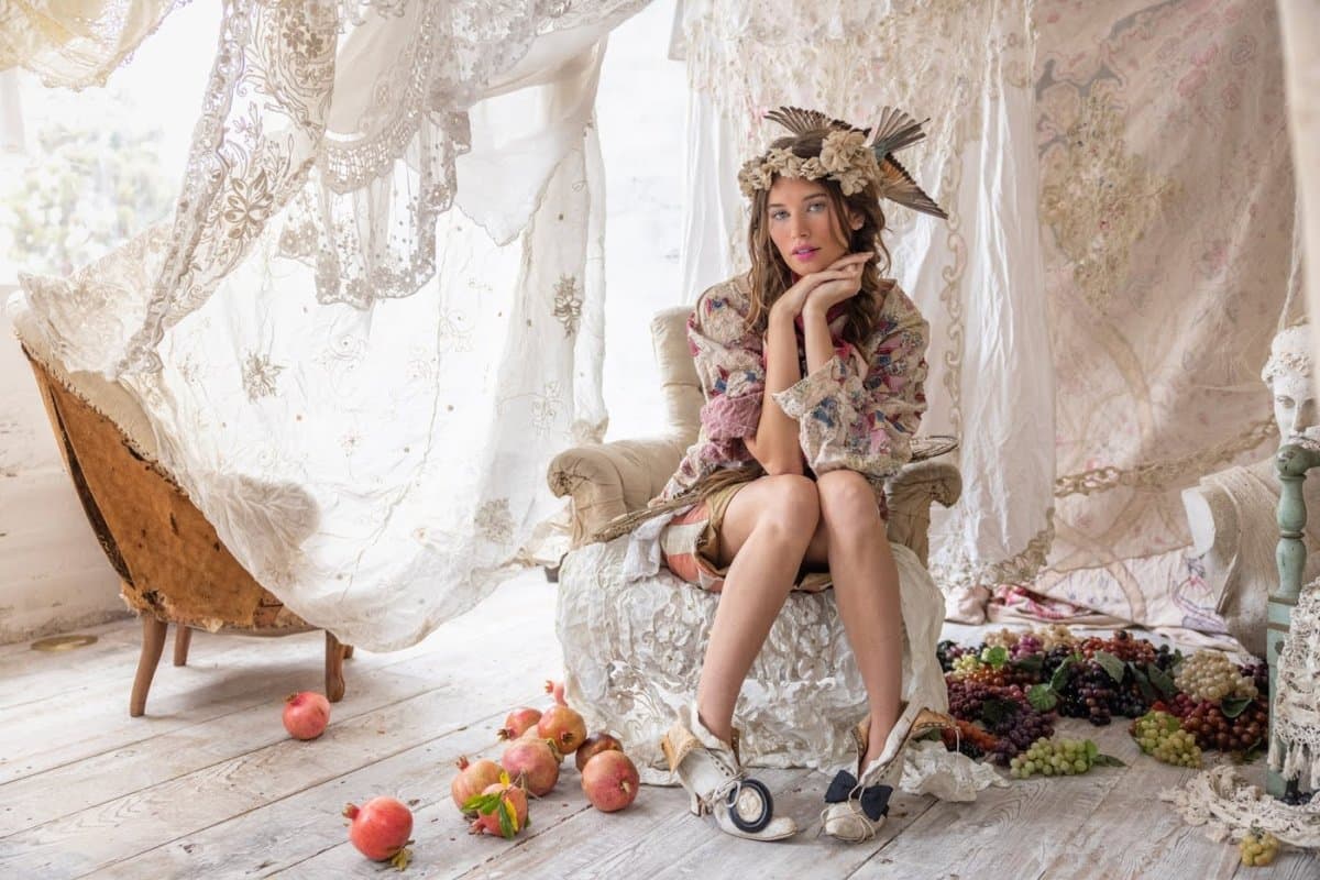 A woman in a floral vintage-style outfit and feathered headpiece sitting among draped lace and scattered pomegranates and grapes