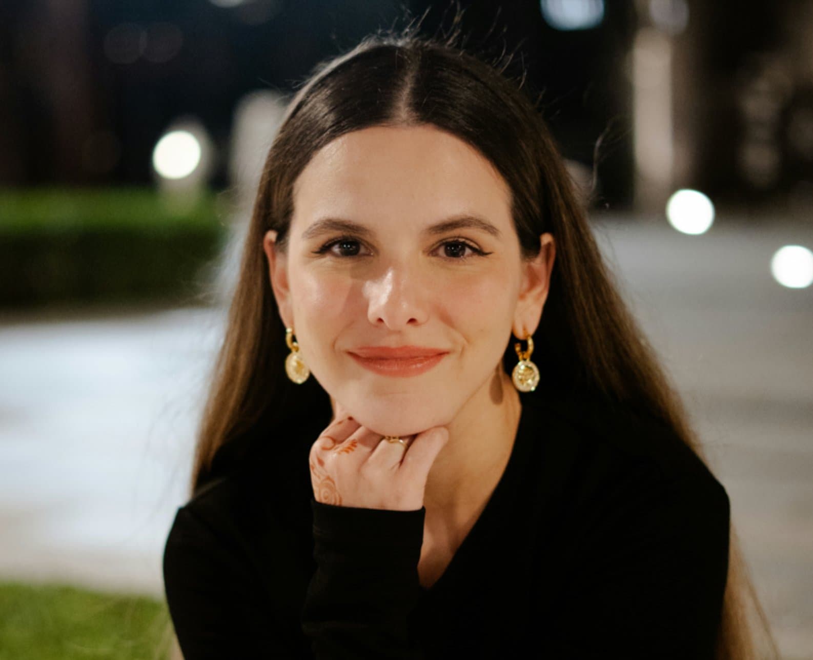 Orianna Zancanaro smiling at night, hand resting under her chin, wearing gold hoop earrings
