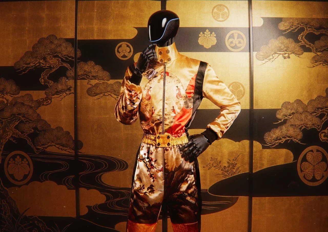 A humanoid robot mannequin wearing a gold satin couture racing suit with hand-painted Japanese-style artwork, posed in front of a gold folding screen