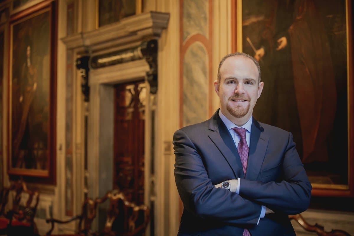 Marco Vidal standing with arms crossed in an ornate gilded room lined with oil paintings