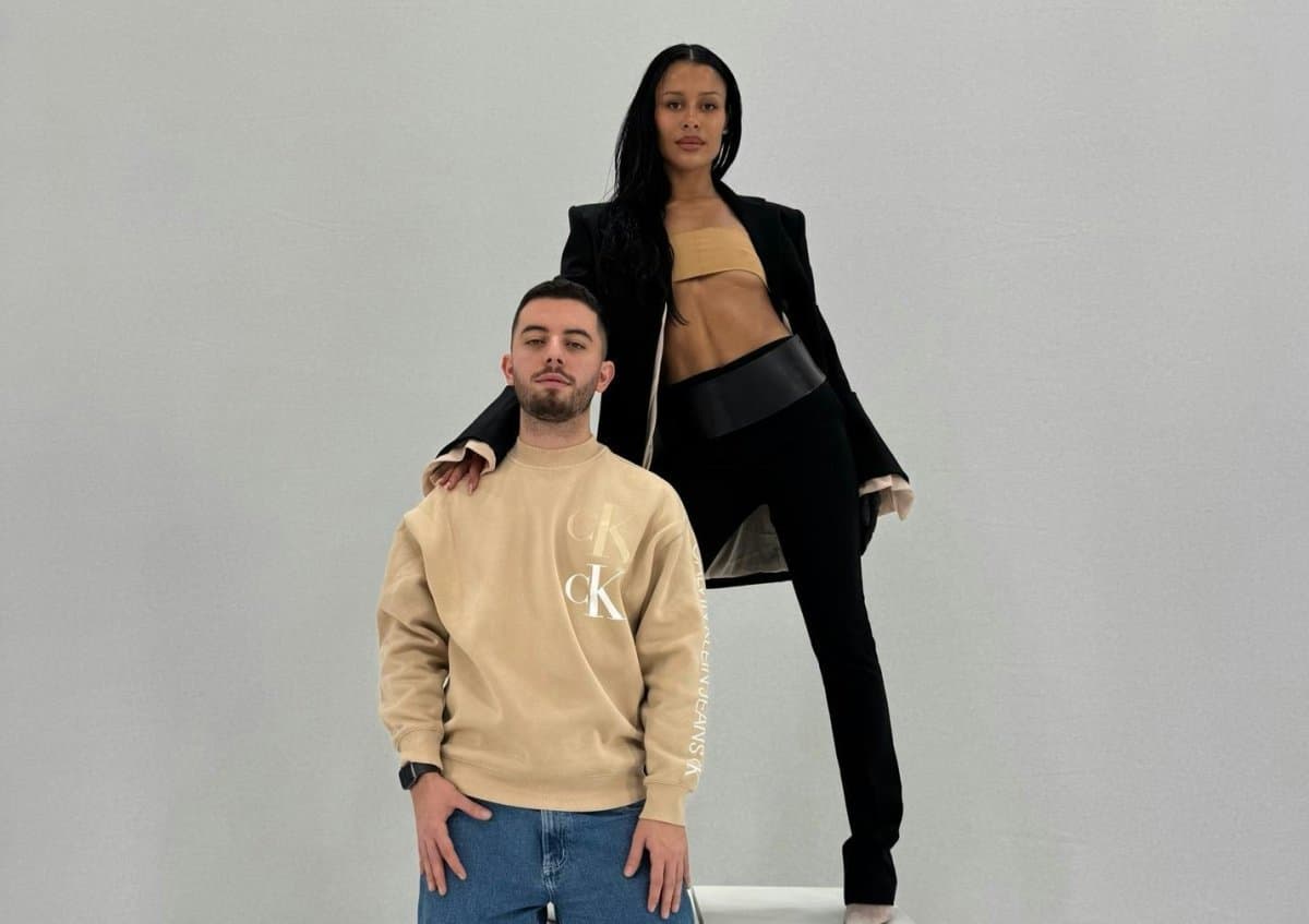 A woman in a black bandeau top and blazer standing behind a seated man wearing a beige Calvin Klein sweatshirt, both against a plain gray backdrop