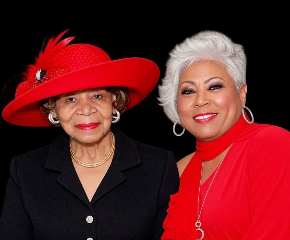 Two women posed for a portrait, one in a black suit and a wide red hat with feather trim, the other in a red dress with silver jewelry