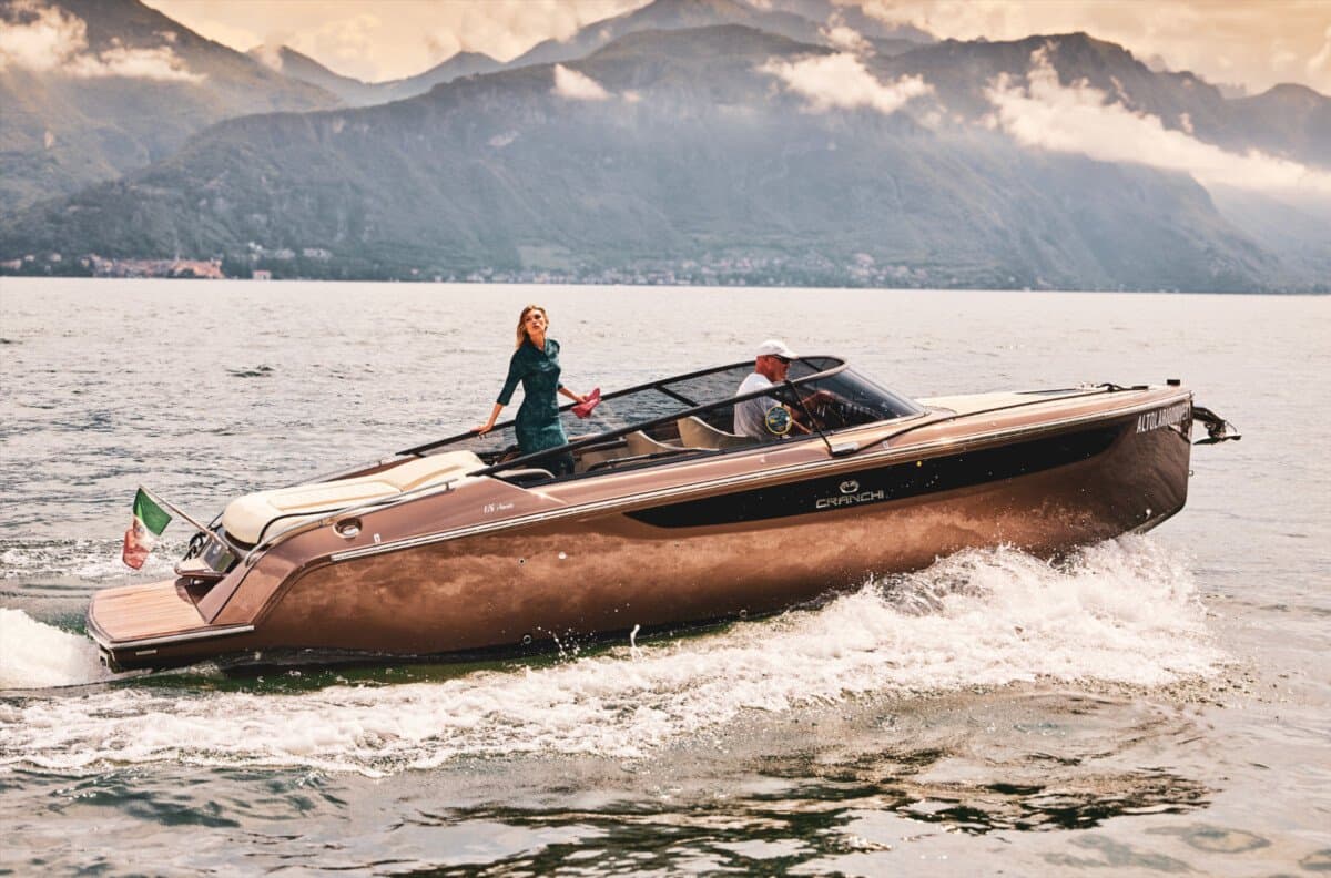 A woman in a green dress standing on a Cranchi motor yacht as it speeds across a lake beneath mountains