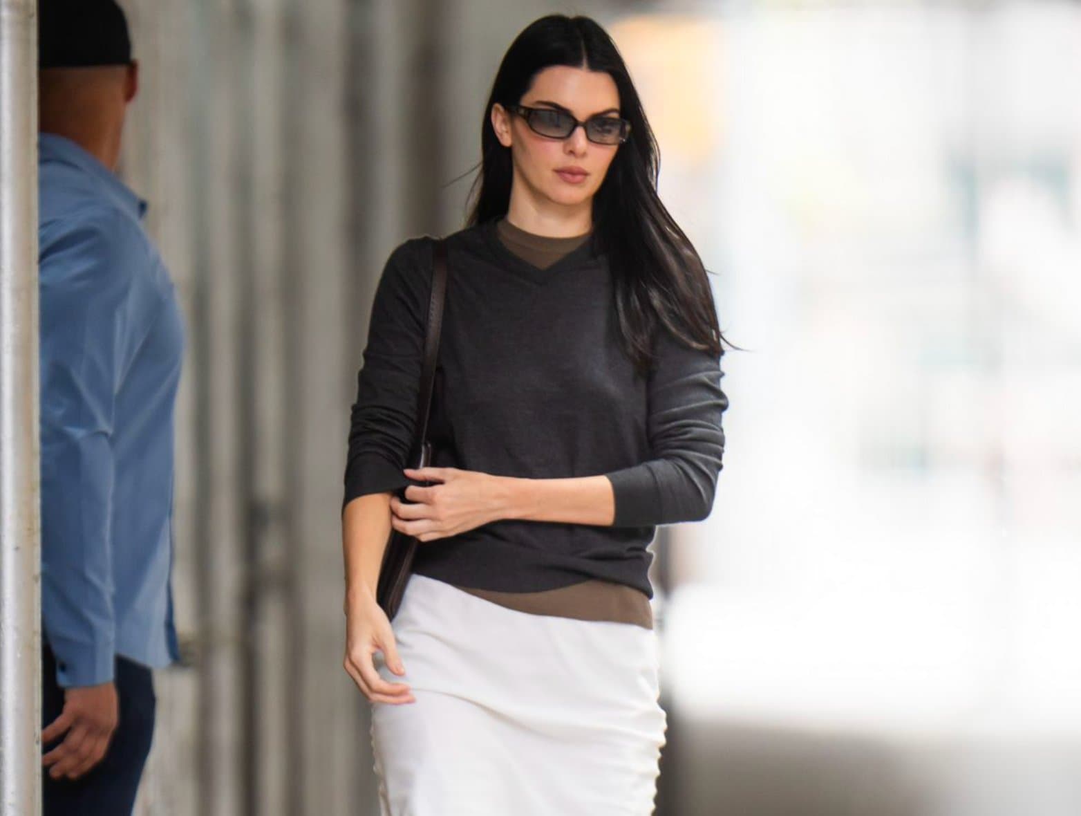A woman in sunglasses wearing a dark sweater over a white satin skirt, walking past a building entrance