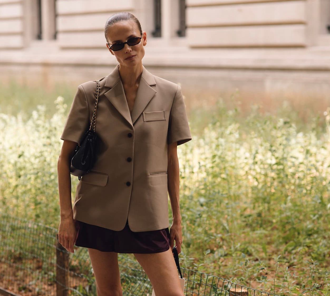 Street style model in a short-sleeve tan blazer, dark sunglasses, and a black shoulder bag, standing in front of a flowering garden