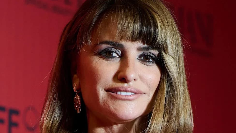 Close-up of Penelope Cruz with a fringed bob hairstyle, gold drop earrings, and smoky eye makeup in front of a red step-and-repeat background