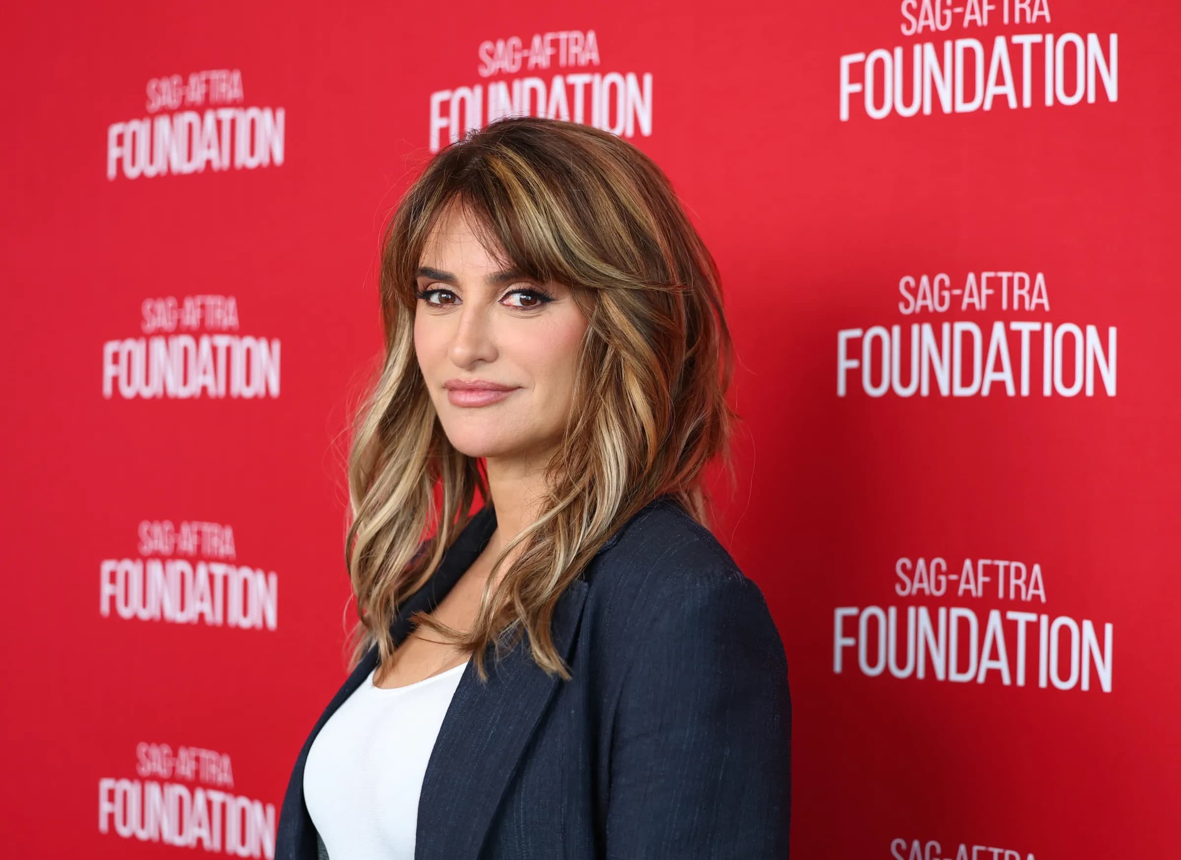 Penelope Cruz at a SAG-AFTRA Foundation event wearing a navy blazer over a white top, in front of a red step-and-repeat background