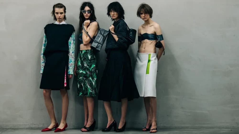Four models backstage at the Prada spring 2027 ready-to-wear show wearing tailored black and printed separates with structured leather bags