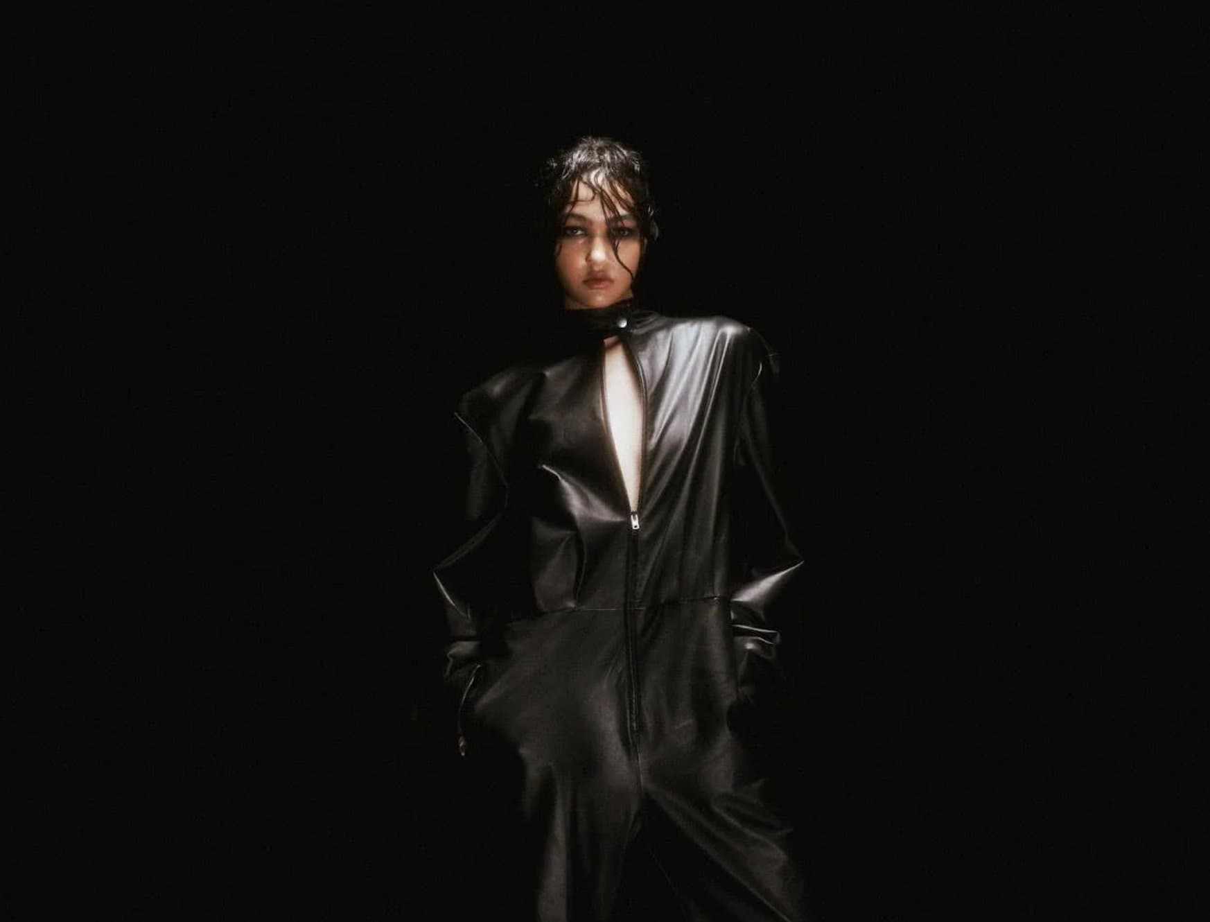 A model on a dark runway wearing a glossy black leather jumpsuit with a keyhole neckline and structured shoulders