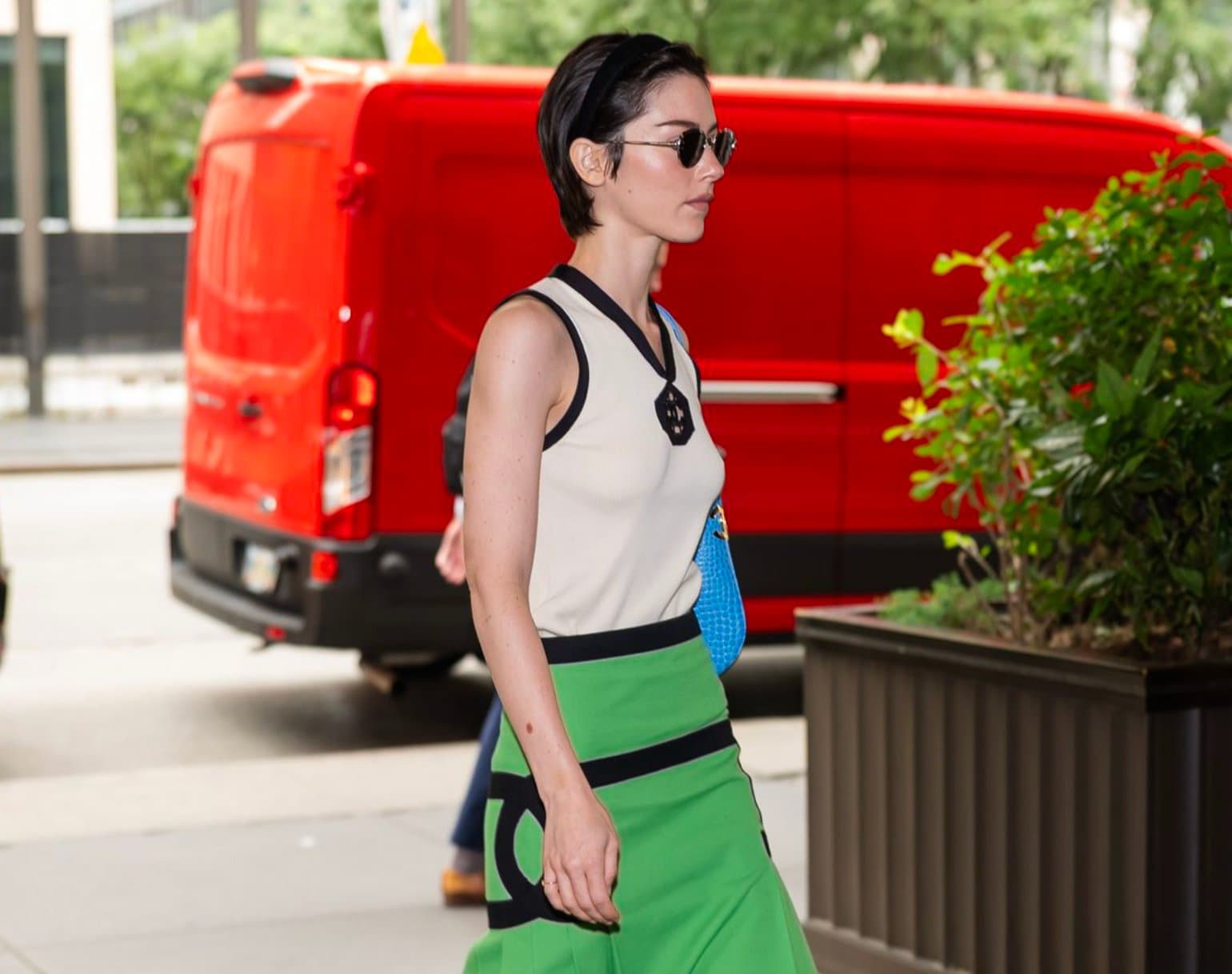 A woman in a black and green color-blocked knit skirt and vest carrying a blue bag, walking past a red van