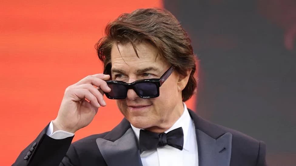 Tom Cruise in a tuxedo and bow tie, adjusting his sunglasses on a red carpet with an orange-lit background