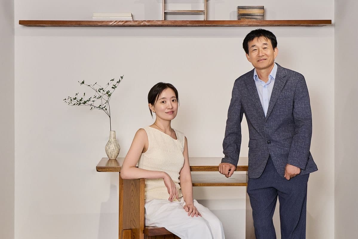 Ceramic artist Baek Gyeong-won seated and woodworker Jo Bok-rae standing beside a handmade wooden desk and shelf displaying a ceramic vase